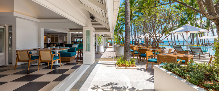Restaurant mit schwarz-weiß kariertem Boden und direktem Zugang zur Terrasse; zahlreiche Tische und Stühle im Fokus, im Hintergrund Palmen und ein leuchtend blauer Pool.